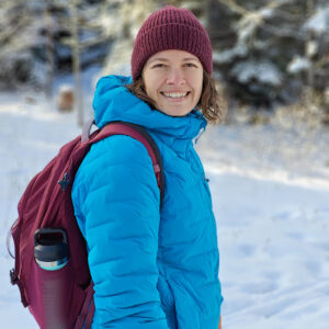 Amy stands out in the snow on a sunny day, trees in background. She wears a plum woolen hat and backpack and a bright blue winter coat