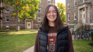 Ruth McGill, wearing a maroon sweatshirt and down vest stands in the Quad with the Bays residences behind her.