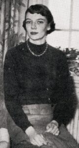 photo from 1956, Ann sits on the arm of a chair. She has a short, dark bob with a window behind her.