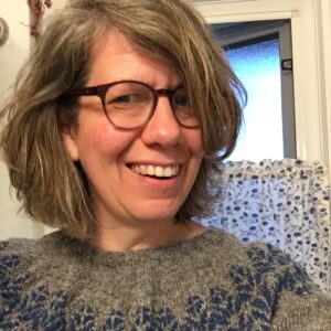 Tracy has shoulder length wavy bob with side part, wearing glasses and smiling wearing a grey sweater with blue detail