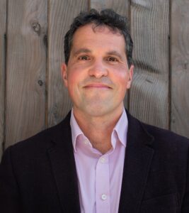 Greg stands in front of a barn board wall wearing a dark shit and button down shirt.