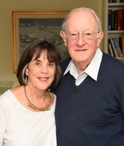 Tia and George Cooper stand arm in arm in front of a painting and bookshelf.