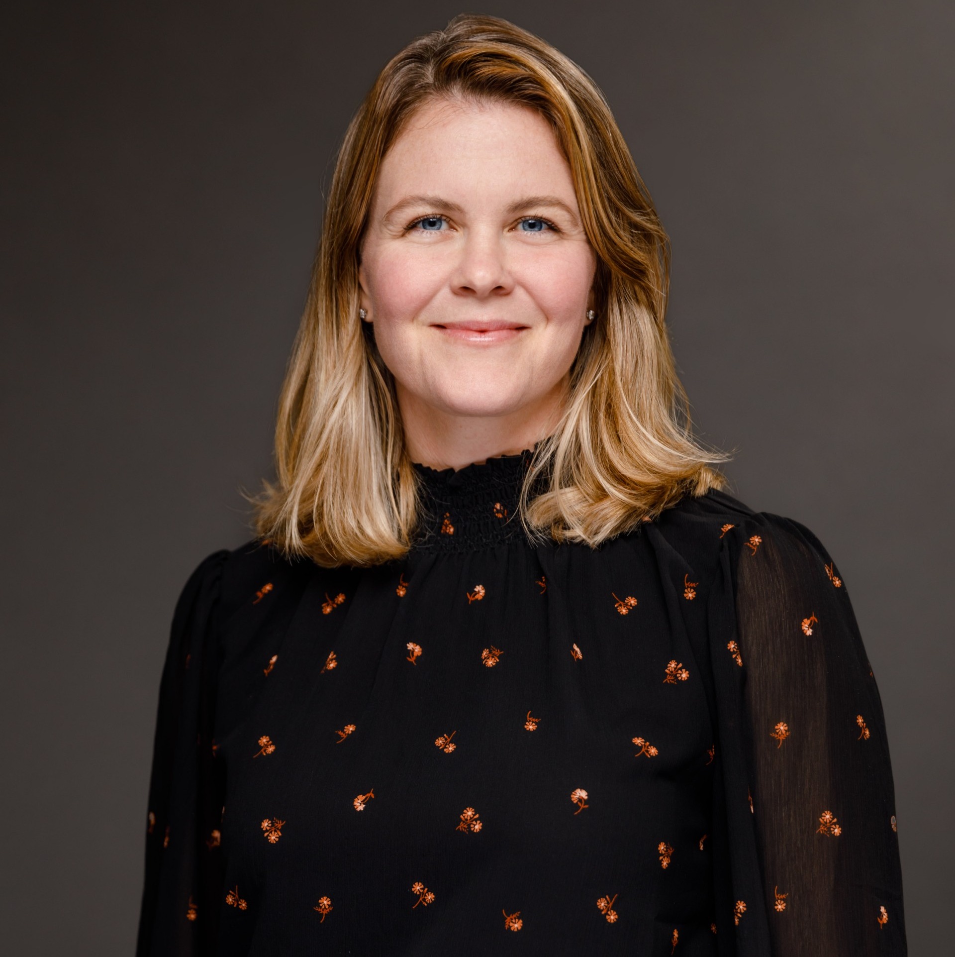 Katherine wears a dark blouse with small flower motif. She stands in front of a neutral backdrop. Has shoulder length blond hair and blue eyes.