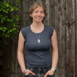 Grad Sam Flood stands outside in front of a wooden fence with vertical slats. She is light skinned with short light brown wavy hair.
