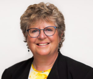 Dr. Stewart has, short, blond hair, wears glasses and dangling earrings. She wears a black jacket with yellow shirt underneath.