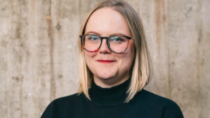 Lyndsie stands near a barnboard wall. She has shoulder length blond hair and wears large round glasses