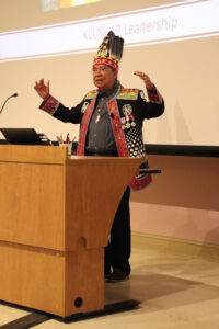 Chief Augustine stands at lectern in regalia, arms raised as he delivers his remarks.