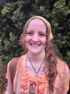 Laura wears an orange shirt with Indigenous symbols. She has her hair tied back with a bandana. She is standing outside in front of an evergreen bush.