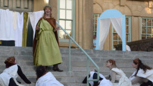 An actor wearing a lime green dress with rusty orange cape and wearing a crown delivers lines as other actors lay at her feet lookin at her