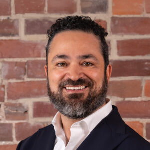 Daniel has dark hair and a beard, wearing a jacket with open shirt collar and is standing in front of a brick wall.