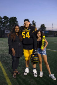 Mateo stands on a football field wearing his uniform. On his right is his mom, and left his sister.