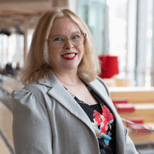 Jennifer stands in modern glass and wooden building atrium. She wears a grey jacket over black shirt. She has shoulder length blond hair and glasses