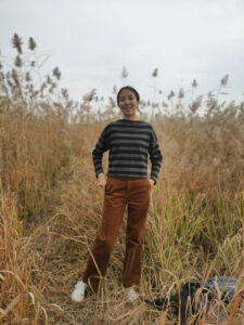 Lu stands in a field surrounded by tall grasses, she has her hands in her pockets and is wearing rusty orange pants and grey long sleeved sweater.