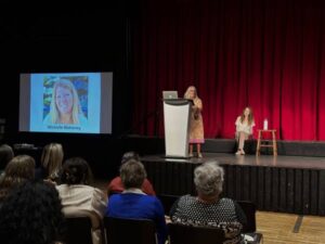 Michelle Mahoney stands on stage behind podium. Her picture is projected on screen to her right. 