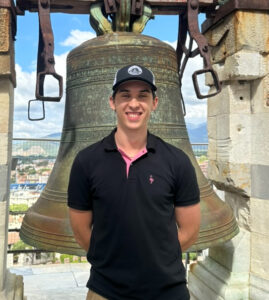 Jai wears a polo shirt and baseball cap. He stands in front of a very old large church bell with the countryside visible in the background.