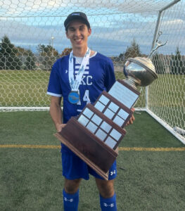 Jai holds the ACAA championship trophy while standing on soccer pitch in his uniform and is sporting a medal.
