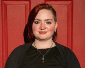 Hailey stands in front of red door. she has dark hair parted on left and is wearing black shirt.