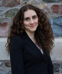 Jenna stands in front of stone wall outside King's. She has long wavy hair and is wearing a dark jacket and looking to camera