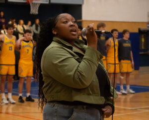 Eriana Willis sings holding microphone in gymnasium before basketball game celebrating African Heritage Night.