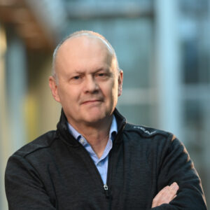 MFA alum Chris Moore wearing dark jacket and blue collared shirt. He is bald, older and has his arms crossed looking at camera.