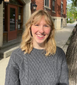 Emily has shoulder-length blond hair and is wearing a grey sweater. She stands outside on a sidewalk with an old red brick building behind her. 