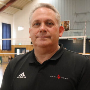 Retired Athletics Director Neil Hooper stands in Gym wearing polo shirt with volleyball net in background