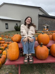 Tessa sits on a red picnic table surrounded by big orange pumpkins. 