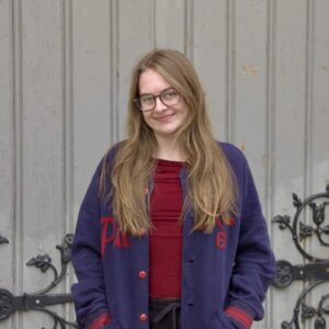Brace wears red shirt under blue and red cardigan. She stands in front of light grey barn wall. She has long fair hair and wears glasses.