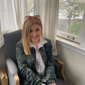 Adriana sits in lounge chair in corner by window. She has long fair hair and has her glasses perched on her head