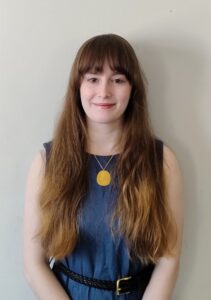 Caroline Jones has long brown hair with bangs and is wearing a belted, blue, sleeveless dress. She is standing against a plain white wall.