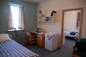 twin bed on left and another visible through a door. In between a desk, chair and small fridge stand on wall separating the two spaces.