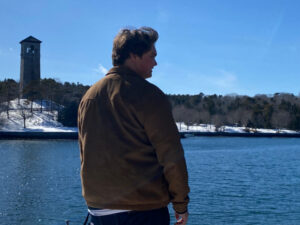 Ethan stands on the shore of the Northwest Arm, Dingle Tower in the background. He stands mostly with his back to camera on a sunny, winter day.