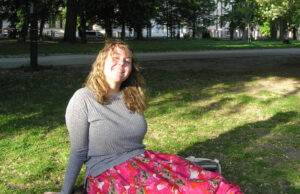 Frances sits on grass leaning back and smiling to camera. She wears a grey sweater and floral pink skirt