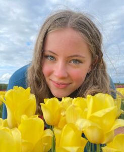 Vivi has long fair hair and blue eyes, she is leaning over a bunch of yellow tulips with the blue sky behind her