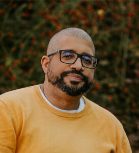 Jon, with bald head, glasses and beard, wears a yellow tshirt outside