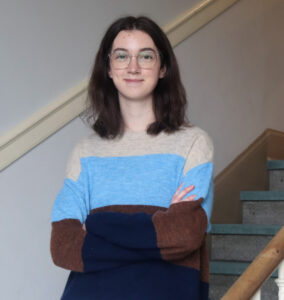 Molly wears a striped sweater and is standing on a staircase. She has long dark hair and wears glasses