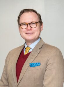Dr. Vine in beige suit with red vest, blue shirt and yellow tie. He wears round glasses and is grinning to camera
