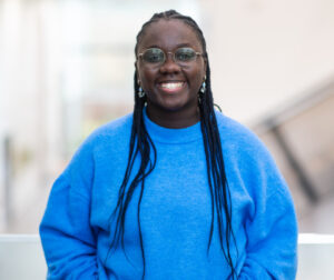 Lucy wears a light blue sweater, long dark braids and has glasses and is smiling to camera