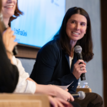 Nina holds a microphone while sitting on stage and is leaning forward to look at someone sitting to her right (not visible.)