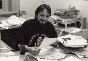 Stephen Kimber at desk surrounded by piles of paper as he holds paper and a pencil. he is looking to camera and smiling